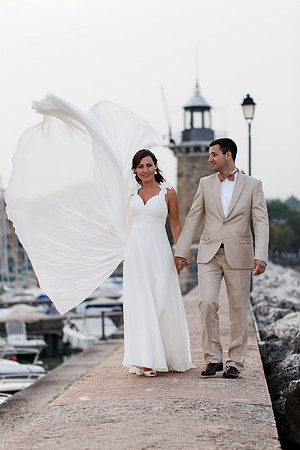 Bride and groom walking by the water, with veil flowing.