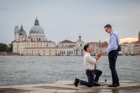 Same-sex couple engagement moment with one man kneeling by the Venice lagoon.