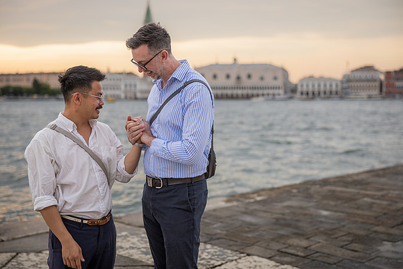 Gay couple holding hands by the water during golden hour in Venice.