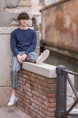 Young man seated by a Venetian canal, posing casually during family photo session.
