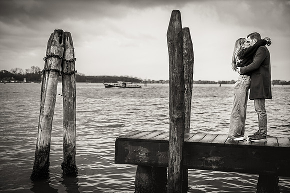 A couple shares an intimate moment by the wooden poles of the Venetian lagoon.