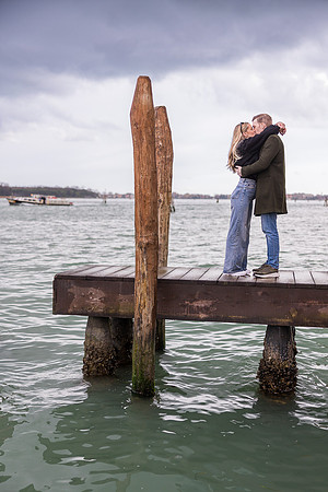 The couple stands on a wooden pier, wrapped in an embrace as the water ripples beneath them.