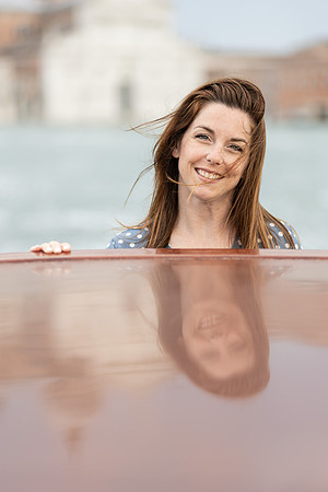 Woman smiling while leaning on a wooden water taxi, her reflection visible.