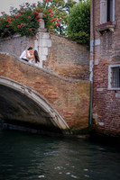 Same-sex couple sitting on a bridge over a Venice canal, surrounded by flowers.