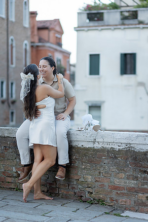 Same-sex couple cuddling on a stone ledge with Venice in the background.