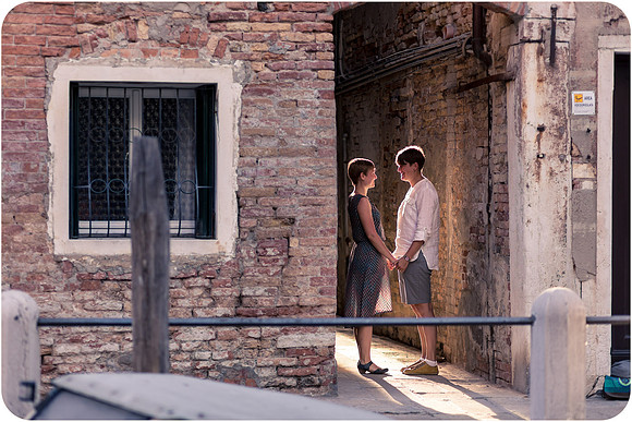 Lesbian couple leaning on a bridge, gazing at the passing gondolas.